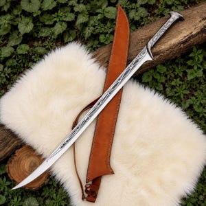 May include: A long, curved metal sword featuring intricate silver-toned filigree engravings along the blade and handle, paired with a tan leather scabbard that includes a thin adjustable strap and visible stitching along the edges.