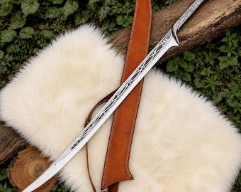 May include: A long, curved metal sword featuring intricate silver-toned filigree engravings along the blade and handle, paired with a tan leather scabbard that includes a thin adjustable strap and visible stitching along the edges.