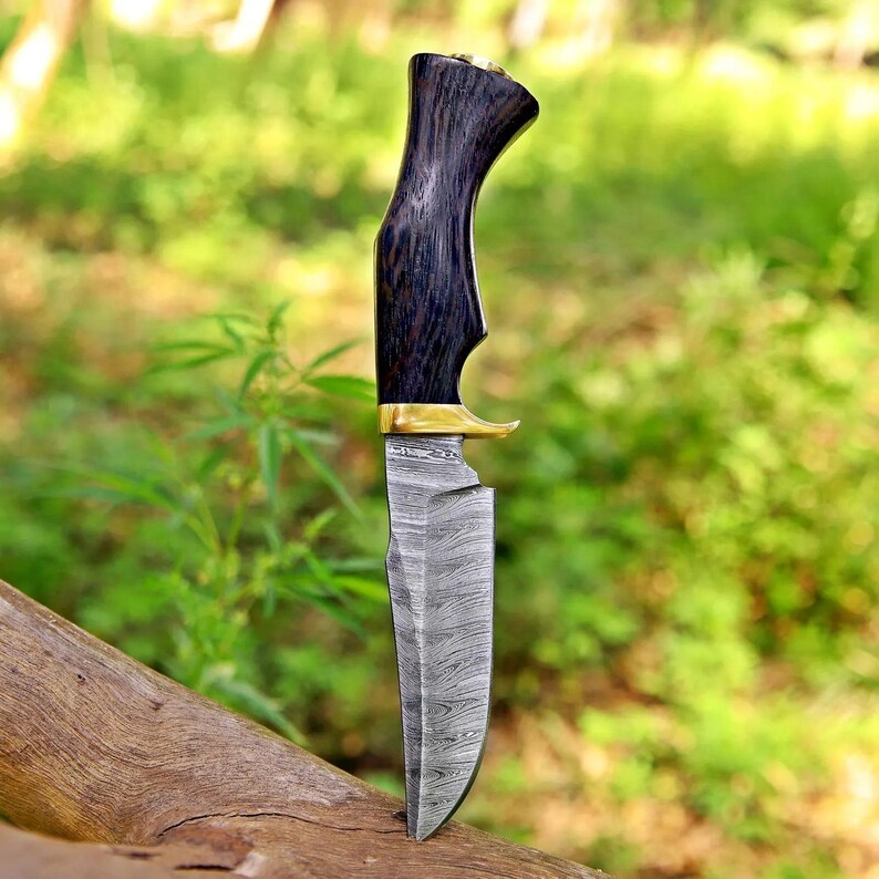 May include: A fixed-blade knife featuring a dark, textured wooden handle with a gold-colored bolster and a silver-toned blade displaying a distinct, wavy, layered metal pattern throughout the entire surface of the steel.