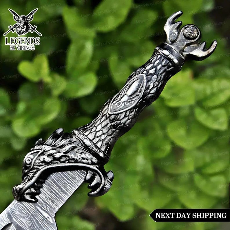 May include: A silver-toned metal dagger handle featuring a detailed dragon head guard, a textured scale-patterned grip with an oval center inlay, and a pommel shaped like a stylized crown. The blade displays a visible layered metal pattern.