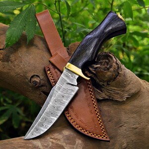 May include: A fixed-blade knife featuring a patterned Damascus steel blade, a dark wood handle with a polished brass bolster, and a tan leather sheath with decorative stitching along the edges and a snap closure strap.