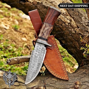 May include: A fixed-blade knife featuring a patterned Damascus steel blade, a bolster, and a handle crafted from a combination of dark wood and textured antler. The knife is accompanied by a tan leather sheath with visible yellow stitching.