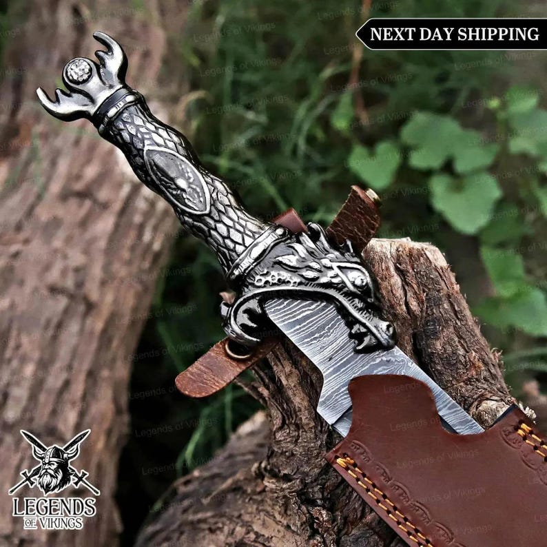 May include: A silver-toned dagger featuring a textured handle with a dragon-shaped crossguard and a patterned blade. The dagger is partially inserted into a brown leather sheath with visible stitching along the edges and a leather strap.