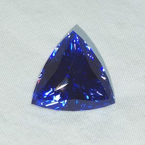 Puede incluir: Una piedra preciosa facetada cortada en forma triangular. La piedra presenta un color azul real profundo y vibrante con reflejos de luz internos y bordes geométricos afilados que crean una apariencia brillante y centelleante.