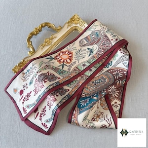 Könnte beinhalten: Ein langer, rechteckiger Seidenschal mit cremefarbenem Hintergrund und einem mehrfarbigen Druck aus Pferden, Blumenmotiven und geometrischen Mustern, eingefasst mit einem dunkelburgunderroten Rand. Der Schal liegt auf einem goldenen Rahmen.