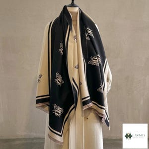 Puede incluir: Una bufanda negra y beige con un patrón repetitivo de abejas y bordes a rayas. La bufanda tiene un dobladillo con flecos y está colocada sobre un maniquí. El logotipo KASHMRA LUXURY STORE es visible en la esquina inferior derecha.