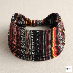 Könnte beinhalten: Ein gemustertes Stirnband in Schwarz, Rot, Gelb und Weiß. Der Stoff weist ein Stammesdesign mit vertikalen Streifen und geometrischen Formen auf. Das Stirnband ist gefaltet und scheint aus einem weichen, flexiblen Material zu bestehen.