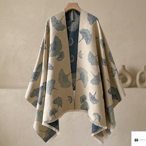 Può includere: Un poncho beige aperto sul davanti con un motivo ripetuto di foglie di ginkgo blu scuro. Il capo presenta una silhouette drappeggiata con una fodera interna blu scuro visibile e un orlo sfrangiato, appeso a una gruccia di legno.