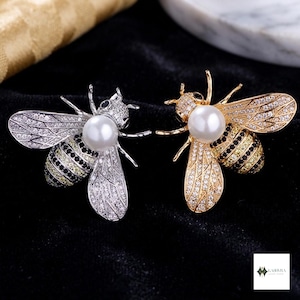 May include: Two bee-shaped brooches, one in silver tone and one in gold tone. Each brooch features a central white pearl, wings encrusted with clear crystals, and a striped abdomen decorated with alternating rows of black and yellow crystals.
