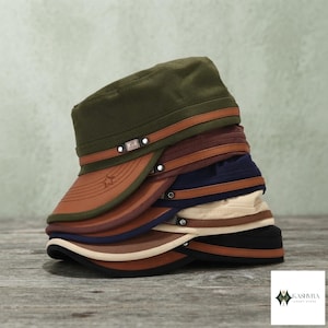 Op de afbeelding: Een stapel van vijf militaire cadet-petten in olijfgroen, bruin, marineblauw, beige en zwart. Elke pet heeft een platte bovenkant, een gebogen klep en een bruine kunstleren band aan de voorzijde met een kleine metalen gesp en een ster.