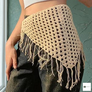 May include: A cream-colored triangular crochet top featuring an openwork granny square pattern and long fringe along the bottom edge. The garment is worn over dark-colored pants and a white top, showcasing the intricate yarn texture and design.