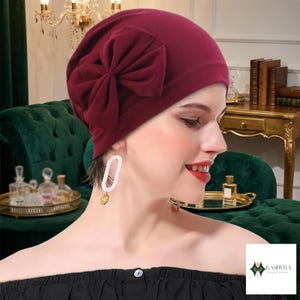 Könnte beinhalten: Eine tiefburgunderrote Mütze mit einer großen, gerafften Stoffschleife an der Seite. Die Mütze besteht aus einem einfarbigen, glatten Stoff und bedeckt den Kopf vollständig, wobei sie eine abgerundete Silhouette mit weichen Falten aufweist.