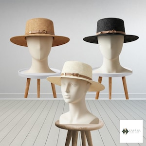May include: Three straw hats in tan, black, and cream, each featuring a flat top, a wide brim, and a thin tan faux leather band with a gold-toned metal buckle detail. The hats are displayed on three separate white mannequin heads on wooden stands.