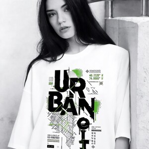 May include: White t-shirt with the word "URBAN CITY" in large black letters. The design includes a map graphic and green accents. The model is wearing the shirt and leaning against a wall.