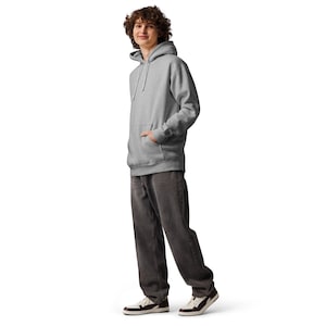 Puede incluir: Una sudadera con capucha gris claro con un bolsillo delantero y una capucha con cordón. La persona lleva vaqueros grises oscuros y zapatillas blancas y marrones. El atuendo es informal y cómodo.