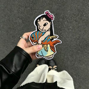 May include: An embroidered patch featuring a cartoon character with black hair and a pink flower, wearing a light blue dress, holding a large orange and blue striped fish. The patch has a white border and is stitched with colorful thread.