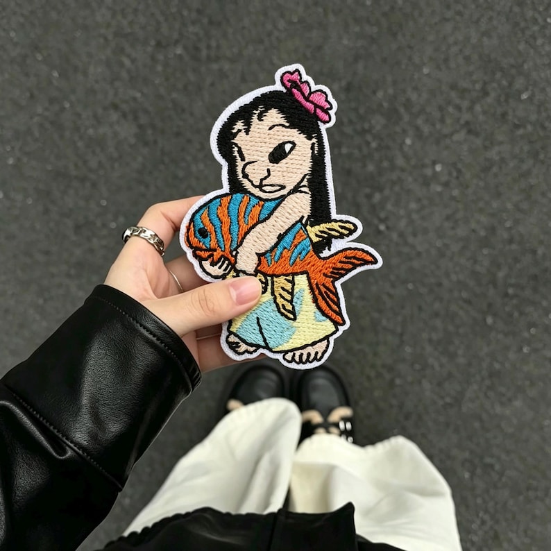 May include: An embroidered patch featuring a cartoon character with black hair and a pink flower, wearing a light blue dress, holding a large orange and blue striped fish. The patch has a white border and is stitched with colorful thread.