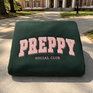 May include: A dark green sweatshirt featuring the word PREPPY in large, pink and white gingham-patterned embroidered letters with a pink outline. Below this, the words SOCIAL CLUB are embroidered in a smaller, simple pink font.