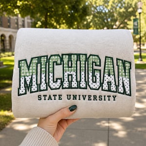 May include: A light beige sweatshirt featuring the text MICHIGAN in large, dark green embroidered letters. The top half of the letters has a green and white gingham pattern, while the bottom half has a white polka dot pattern. STATE UNIVERSITY is printed below.