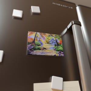 May include: A rectangular magnet featuring a colorful landscape painting of a river scene with two animals. The artwork includes trees, flowers, and a stream, with a vibrant display of colors. The magnet is attached to a dark brown refrigerator.