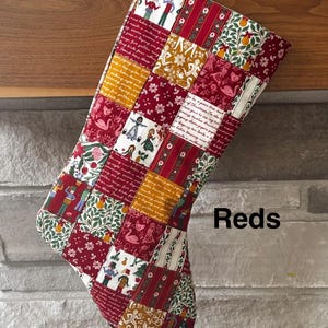 May include: A patchwork Christmas stocking featuring a variety of fabric squares with red, white, and gold tones. The patterns include illustrations of figures, birds, pears, and cursive text. The word Reds is printed in black on the right side.