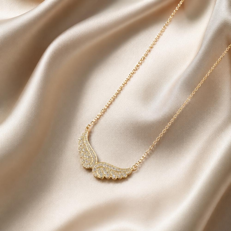 May include: A gold-colored necklace with a pendant shaped like two wings. The wings are encrusted with small, sparkling crystals. The necklace chain is delicate, and the background is a satin fabric.