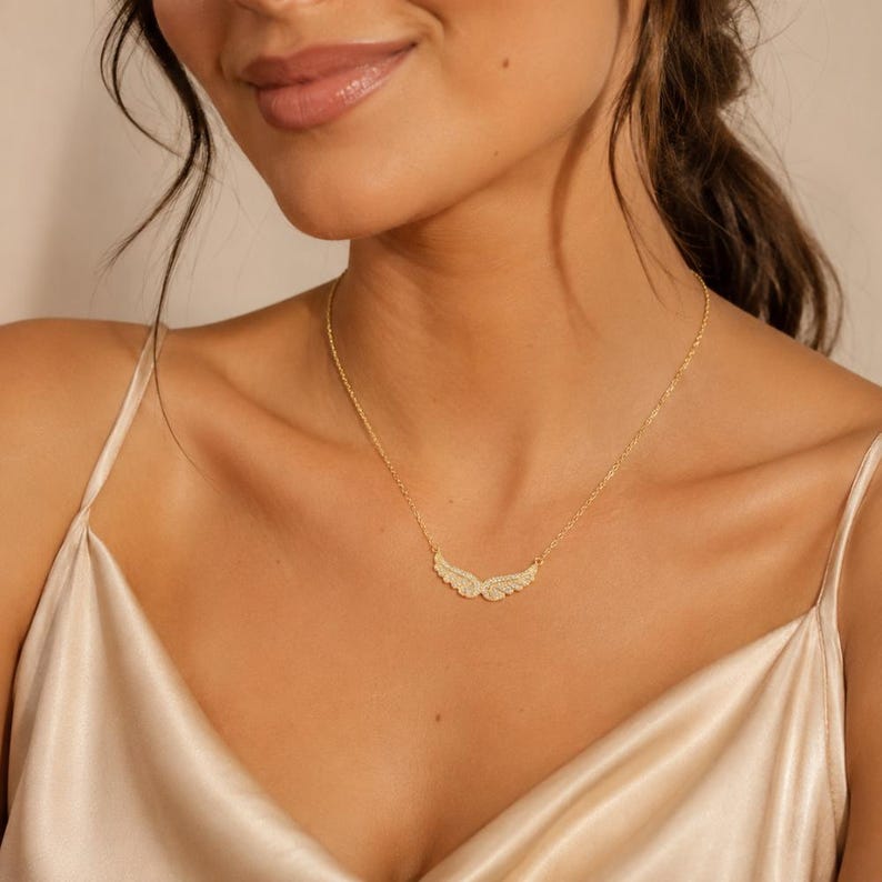 May include: A gold-colored necklace with a pendant shaped like angel wings. The wings are encrusted with small, sparkling stones. The necklace is worn on a person with fair skin and a cream-colored top.