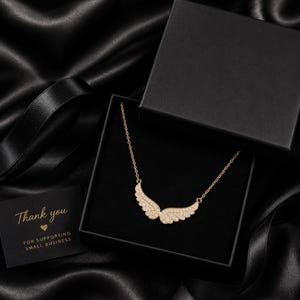May include: A gold-colored necklace with a wing design, encrusted with small, sparkling stones, is displayed in an open black gift box. A small card with the words "Thank you" is also in the box. The background is black.