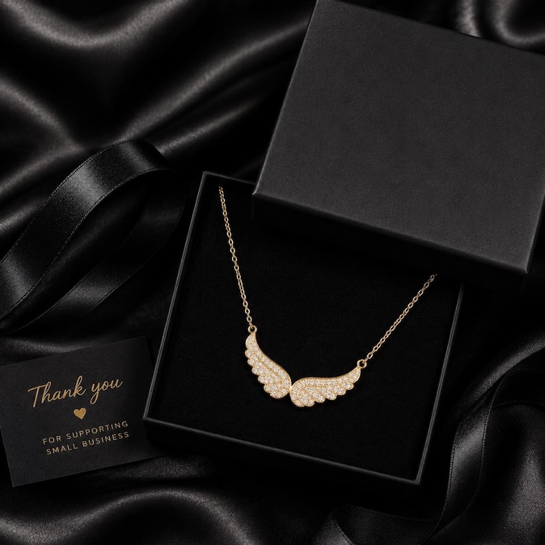May include: A gold-colored necklace with a wing design, encrusted with small, sparkling stones, is displayed in an open black gift box. A small card with the words "Thank you" is also in the box. The background is black.