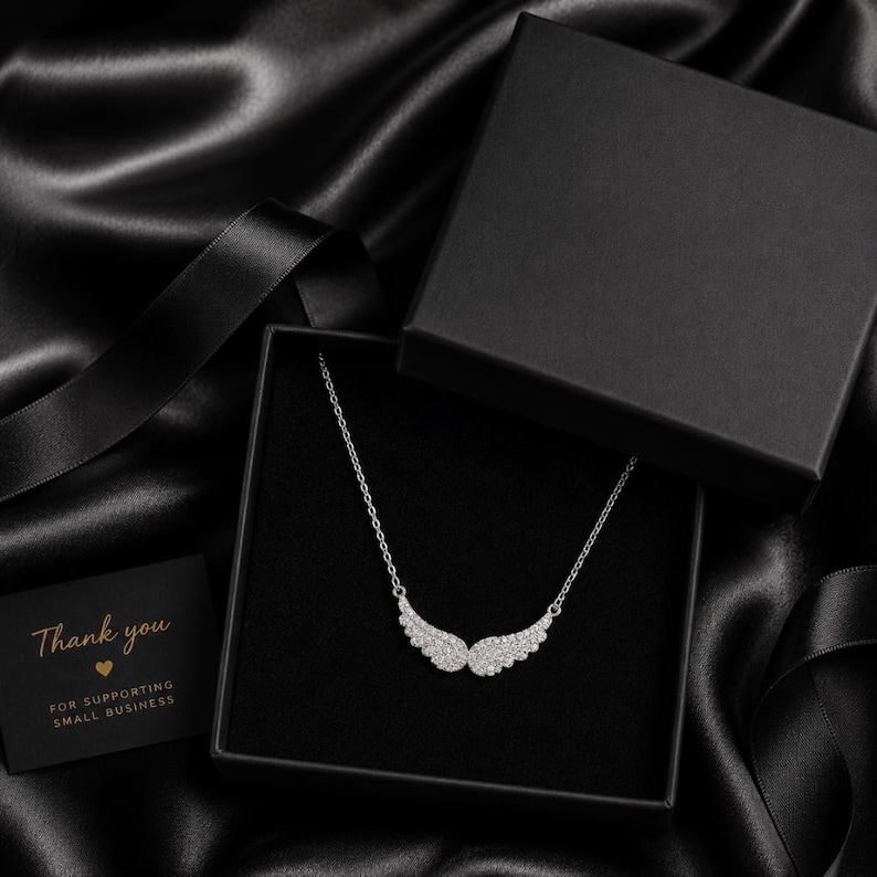 May include: A silver necklace with a wing pendant is displayed in a black gift box. The box sits on a black satin surface. A small card with the words "Thank you" is also present.