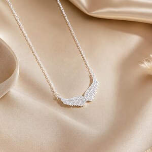 May include: A silver necklace featuring a pair of stylized wings, encrusted with sparkling crystals. The wings are positioned at the center of a delicate chain, creating a graceful and elegant design. The necklace is displayed on a satin surface.