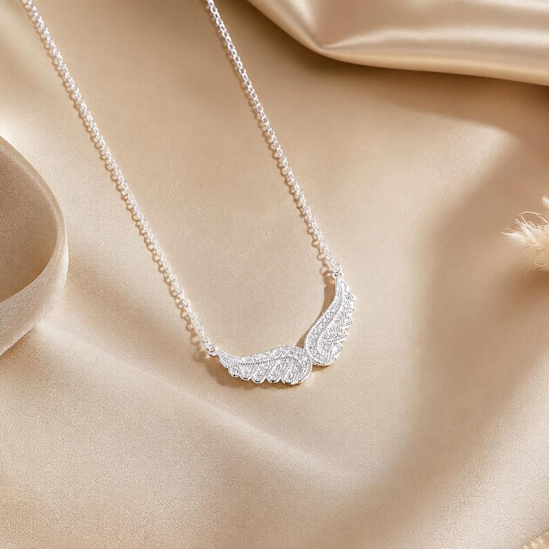 May include: A silver necklace featuring a pair of stylized wings, encrusted with sparkling crystals. The wings are positioned at the center of a delicate chain, creating a graceful and elegant design. The necklace is displayed on a satin surface.