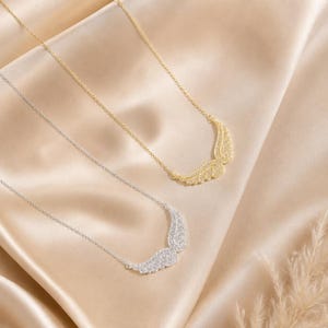 May include: Two necklaces with wing pendants. One necklace is gold-toned, and the other is silver-toned. Both necklaces have a delicate chain and are set against a satin background.