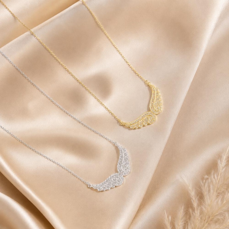 May include: Two necklaces with wing pendants. One necklace is gold-toned, and the other is silver-toned. Both necklaces have a delicate chain and are set against a satin background.