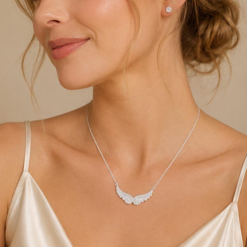 May include: A silver necklace with a pendant shaped like angel wings, encrusted with small, sparkling stones. The necklace is worn on a person with a white satin top. A matching stud earring is visible.
