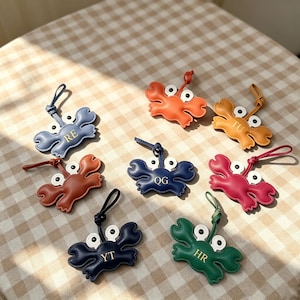 May include: A collection of eight crab-shaped luggage tags in various colors including blue, orange, yellow, brown, dark blue, red, and green. Each tag features two white eyes with black pupils and a gold-stamped two-letter monogram on the center.
