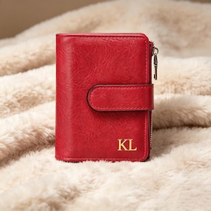 May include: A vibrant red bi-fold wallet with a textured finish, featuring a snap-button closure strap and a side zip with a metal pull. The front of the wallet is embossed with the gold-coloured capital letters KL in the bottom right corner.