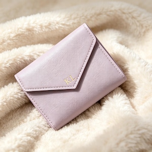 May include: A light lavender envelope-style wallet with a smooth, textured finish. The front flap features the gold-colored embossed letters KL centered on the point. The wallet has a structured, folded design with visible stitching along the edges.