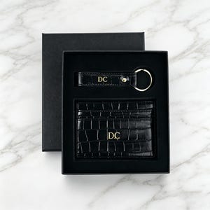 Può includere: Un portacarte nero con stampa coccodrillo e un portachiavi abbinato con anello dorato, entrambi caratterizzati dalle iniziali DC impresse in oro. Il set è presentato in una scatola regalo nera con coperchio rimovibile e inserto interno su misura.