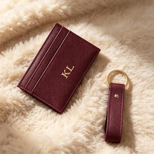 May include: A burgundy card holder with a textured finish and the gold embossed letters KL, accompanied by a matching burgundy keychain featuring a gold metal ring and a small gold stud detail, both displaying a consistent crosshatch pattern.