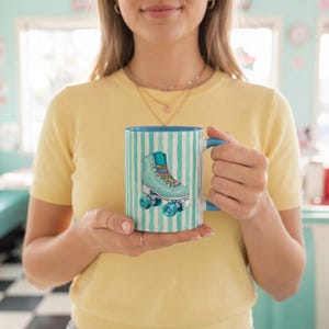 May include: A ceramic mug featuring a light blue and white vertical striped pattern with a central illustration of a light blue roller skate with multi-colored laces and blue wheels. The mug has a solid blue handle and a matching blue interior.