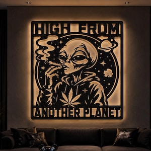 Op de afbeelding: Een vierkant zwart metalen wandobject met achtergrondverlichting, voorzien van een alien in een hoodie met een cannabisbladontwerp. De alien rookt en is omringd door sterren en planeten. De tekst HIGH FROM ANOTHER PLANET is in het frame gesneden.