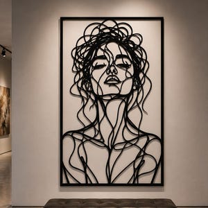 Puede incluir: Una gran pieza de arte mural rectangular que presenta un dibujo de línea continua negra del rostro y los hombros de una persona. La obra de arte está encerrada en un marco negro delgado y montada en una pared blanca con líneas fluidas.