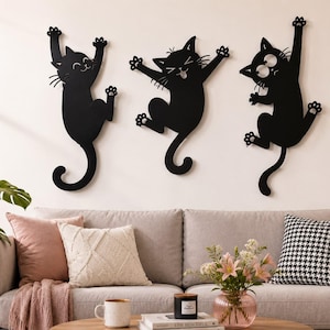 Puede incluir: Tres siluetas de arte mural en metal negro que representan gatos en varias poses de escalada y salto. Cada figura de gato presenta expresiones faciales, bigotes y detalles de patas distintos, diseñadas para montarse en una superficie.
