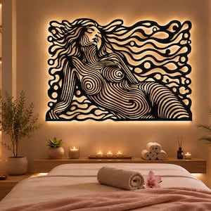 Puede incluir: Una pieza de arte mural negra que presenta una figura reclinada estilizada compuesta por líneas fluidas e intrincadas. El diseño está retroiluminado, creando un brillo cálido alrededor de los bordes del recorte, contrastando con el patrón lineal oscuro.