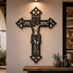 May include: Black metal wall art depicting Jesus on the cross, with the letters INRI at the top. The cross has ornate, decorative edges. The artwork is mounted on a light-colored wall, with a wooden table and decorative items in the foreground.