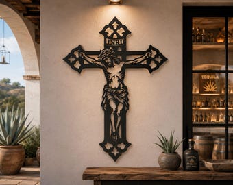Puede incluir: Arte mural de metal negro que representa a Jesús en la cruz, con las letras INRI en la parte superior. La cruz tiene bordes ornamentados y decorativos. La obra de arte está montada en una pared de color claro, con una mesa de madera y objetos decorativos en primer plano.