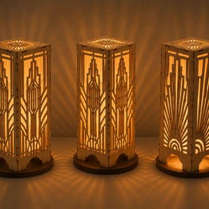 May include: Three rectangular wooden table lamps with intricate laser-cut geometric patterns. Each lamp features a light-colored wood frame with cutouts that emit a warm, glowing light, casting decorative shadows on the surrounding surface.