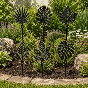 Puede incluir: Un conjunto de seis estacas de jardín de metal negro con diversas formas de hojas, incluyendo una palmera abanico, monstera y helecho. Las estacas están colocadas en un parterre con follaje verde y flores de fondo. Diseñadas para decorar espacios exteriores.