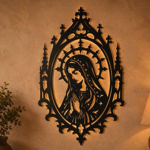 May include: A black laser-cut wall hanging featuring a religious figure in an oval frame. The artwork shows a detailed silhouette of a woman with a halo, hands clasped in prayer, mounted on a beige wall. The frame has gothic-style detailing.